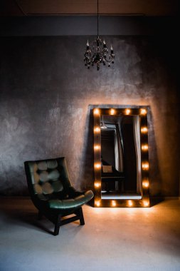 yeşil deri koltuk ve uzun giyinme aynası. Koyu renkli bir odada, gri bir duvarın arka planında, üzerinde lambalar olan bir çerçeve.