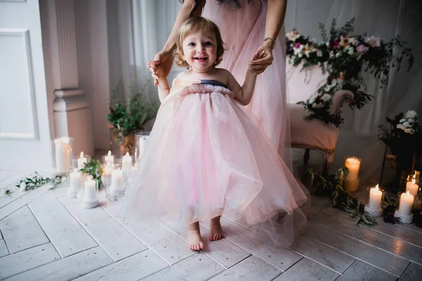 Anne küçük bir kız çocuğu tutuyor, ilk adımlarını atıyor, pembe güzel elbiseler içinde, çiçeklerle ve mumlarla süslenmiş beyaz bir stüdyo odasında, mutlu güzel bir fotoğraf.