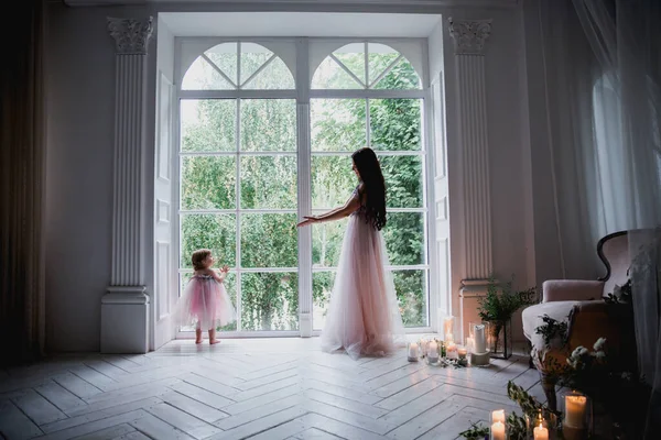Anne ve kızı büyük beyaz bir pencerenin yanında güzel pembe şifon elbiseler içinde klasik dekore edilmiş bir odada duruyorlar.