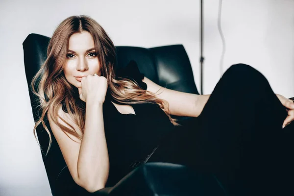 Siyah giysili güzel sarışın kadın portresi beyaz arka plandaki sandalyede oturuyor ve saçları uzayıp gülümsüyor.