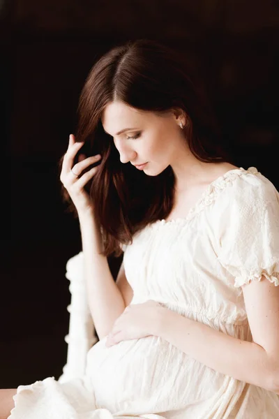 美しい妊娠中のブルネットの女性の白いドレス暗い背景の上にベッドの上に座って — ストック写真