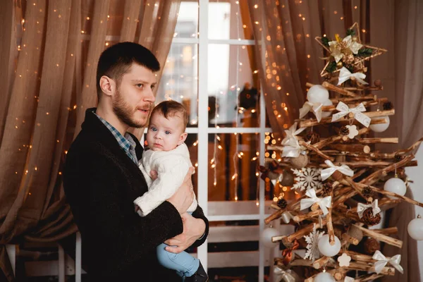 Mutlu bir baba, kollarında oğlu, şeffaf bir perdesi, ışıkları ve Noel ağacı olan bir pencerenin arkasında.