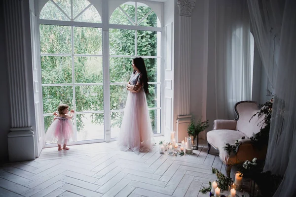 Anne ve kızı büyük beyaz bir pencerenin yanında güzel pembe şifon elbiseler içinde klasik dekore edilmiş bir odada duruyorlar.