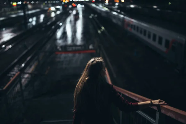 美しい少女が階段の上に立っている 駅の裏側では街灯が通りの夜に水溜りに反射して雨が降り列車が横に見えるのを待っています — ストック写真