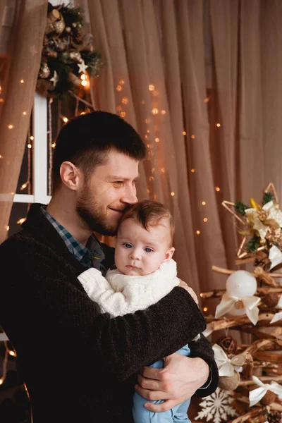 Mutlu bir baba, kollarında oğlu, şeffaf bir perdesi, ışıkları ve Noel ağacı olan bir pencerenin arkasında.