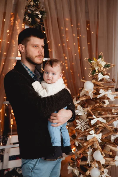 Mutlu bir baba, kollarında oğlu, şeffaf bir perdesi, ışıkları ve Noel ağacı olan bir pencerenin arkasında.