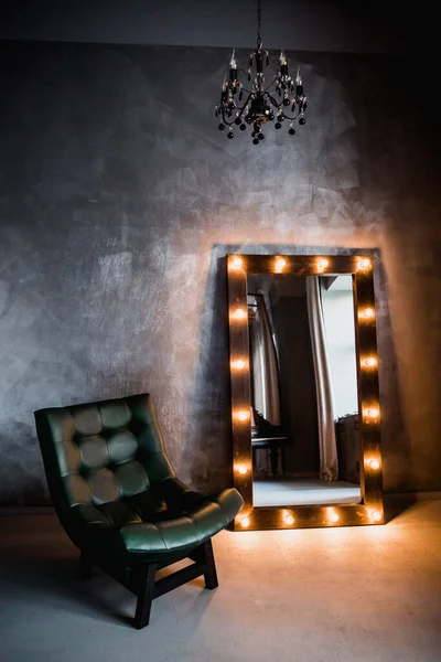 yeşil deri koltuk ve uzun giyinme aynası. Koyu renkli bir odada, gri bir duvarın arka planında, üzerinde lambalar olan bir çerçeve.
