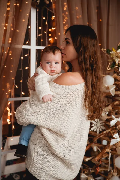 Mutlu anne, oğlunun kollarında rahat kazaklar içinde şeffaf bir perdesi, ışıkları ve Noel ağacı olan bir pencerenin arkasında duruyor.