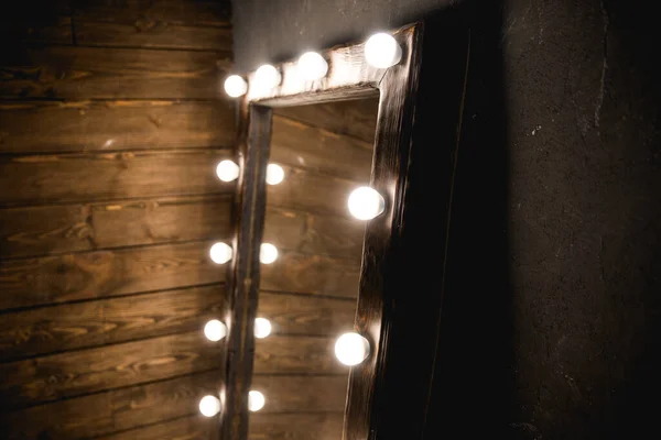 Long dressing mirror with light bulbs stands on the floor against a wooden and concrete wall in loft style room