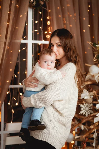 Mutlu anne, oğlunun kollarında rahat kazaklar içinde şeffaf bir perdesi, ışıkları ve Noel ağacı olan bir pencerenin arkasında duruyor.