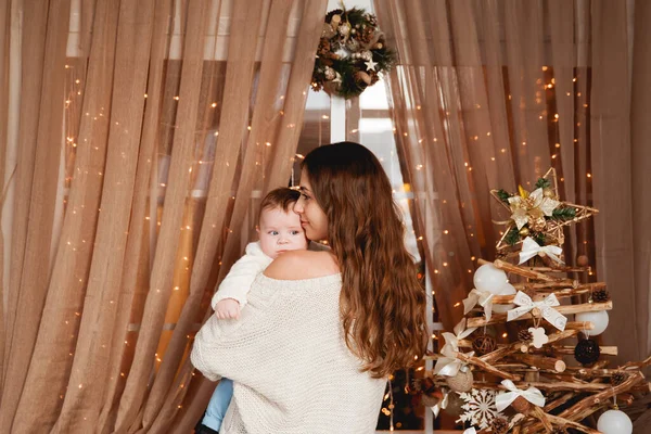 Mutlu anne, oğlunun kollarında rahat kazaklar içinde şeffaf bir perdesi, ışıkları ve Noel ağacı olan bir pencerenin arkasında duruyor.