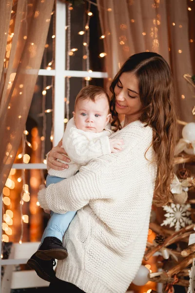 Mutlu anne, oğlunun kollarında rahat kazaklar içinde şeffaf bir perdesi, ışıkları ve Noel ağacı olan bir pencerenin arkasında duruyor.