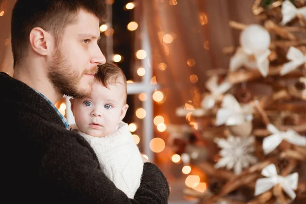 Mutlu bir baba, kollarında oğlu, şeffaf bir perdesi, ışıkları ve Noel ağacı olan bir pencerenin arkasında.