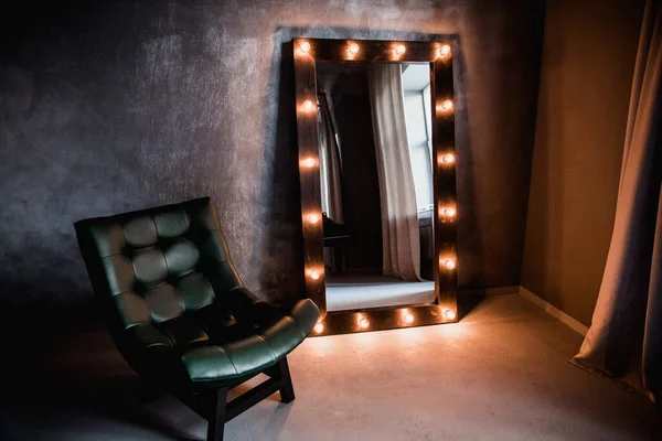 yeşil deri koltuk ve uzun giyinme aynası. Koyu renkli bir odada, gri bir duvarın arka planında, üzerinde lambalar olan bir çerçeve.
