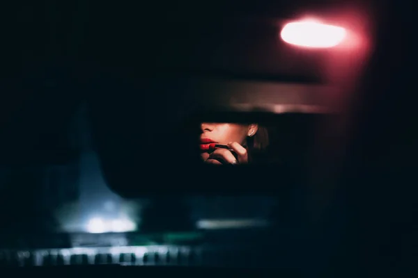 Güzel bir kadının modern fotoğrafı dudaklarına kırmızı bir ruj sürerken gece arabasının dikiz aynası kullanırken çekilmiş.
