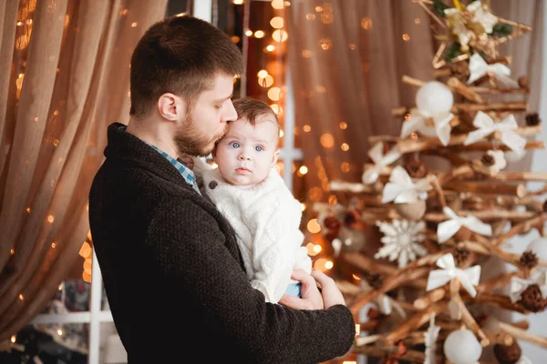 Mutlu bir baba, kollarında oğlu, şeffaf bir perdesi, ışıkları ve Noel ağacı olan bir pencerenin arkasında.