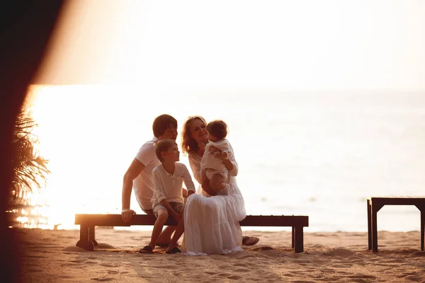 Young happy family in white clothes sits on the beach at sunset