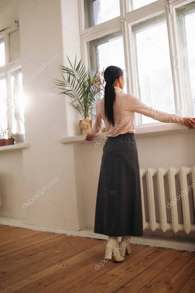 stylish young woman stay near large window in room with wooden floor and white wall 