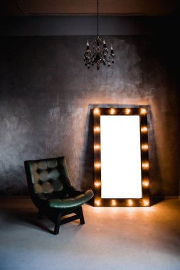 yeşil deri koltuk ve uzun giyinme aynası. Koyu renkli bir odada, gri bir duvarın arka planında, üzerinde lambalar olan bir çerçeve.