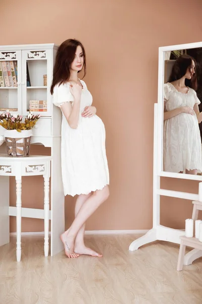 Όμορφη Έγκυος Μελαχρινή Γυναίκα Λευκό Φόρεμα Κοιτάζει Ένα Λευκό Καθρέφτη — Φωτογραφία Αρχείου