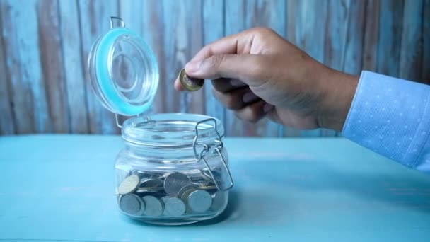 Człowiek wlewa monety do szklanego słoika na stole. — Wideo stockowe