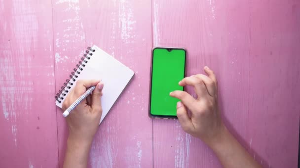 Vista superior de la mano de las mujeres El uso de teléfono inteligente y la escritura en el bloc de notas — Vídeos de Stock