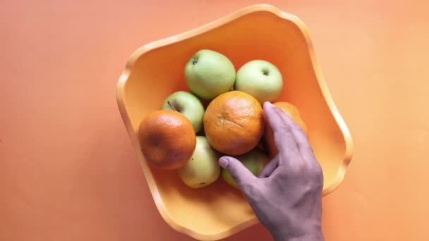 Vista superior de la mano recogiendo fruta naranja de un tazón sobre fondo naranja . — Vídeos de Stock