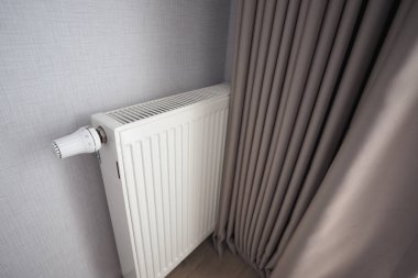 Odada yumuşak gri bir perdenin yanında beyaz bir radyatör var..