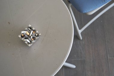 Sigara izmaritleriyle dolu bir kül tablası bir sandalyenin yanında oturuyor..