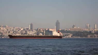 Bir kargo gemisi gün batımında hareketli bir şehir arkaplanıyla denizde yol alır..