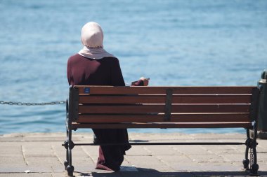 Bir kadın tesettüre oturmuş, su kenarında huzurlu bir anın tadını çıkarıyor..