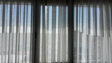 Güneş ışığı, beyaz perdelerin arasından odaya doluyor ve şehir manzarasını ortaya çıkarıyor..