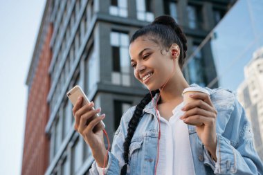 Güzel Afrikalı Amerikalı kadın cep telefonu kullanıyor, iletişim kuruyor, dijital ekrana bakıyor, sokakta yürüyor. Havalı hippi kız elinde akıllı telefon ve bir fincan kahveyle müzik dinliyor.