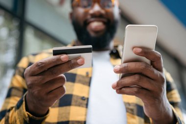 Kredi kartı ve cep telefonu tutan, internetten alışveriş yapan adamların yakınına. Mutlu Afrikalı Amerikalı serbest çalışan ödemeyi aldı, ellerine odaklandı