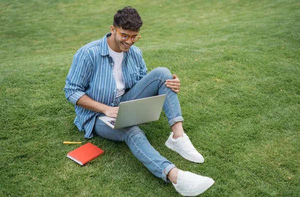 Happy asian student studying, learning language, online education, sitting on grass. Young handsome Indian man using laptop computer, working outdoors, watching training courses