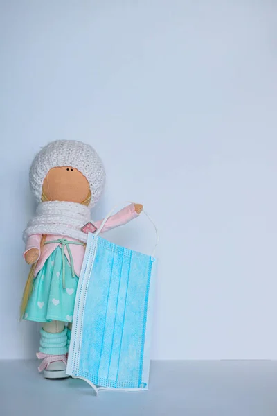 A textile doll in mint pink clothes and a white knitted hat holds a protective face medical mask. COVID-19 coronavirus. Space for text.