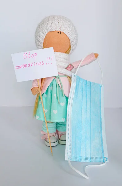 A textile doll in mint pink clothes and a white knitted hat holds a protective medical mask and a sign that reads \