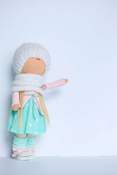 A textile doll in mint pink clothes and a white knitted hat points her hand at text that can be added. There is a clean space for text.