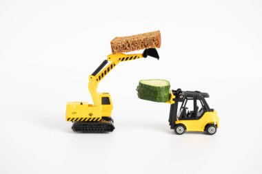 Oyuncak yükleyici ya da parlak sarı renkli inşaat vinci bir parça salatalık ve taze pişmiş bir parça siyah ekmek taşır. Beyaz arkaplanda yakın plan maketler.