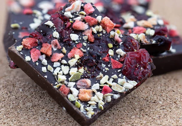 Mantar ahşap arka planda, böğürtlenli ve yeşil fıstıklı el yapımı acı çikolata. Yiyeceğin makro fotoğrafçılığı. Kırık bir çikolatanın yakın çekimi..