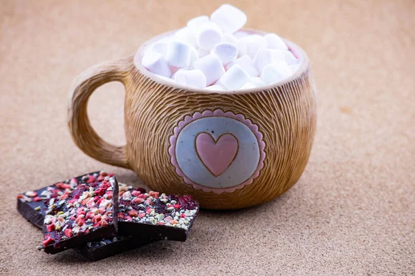 Pembe kalpli kahverengi seramik bardak. Bir fincan kahve veya çay havadar marşmelovlarla doldurulur. Yakınlarda böğürtlen ve şam fıstıklı acı siyah çikolata var..