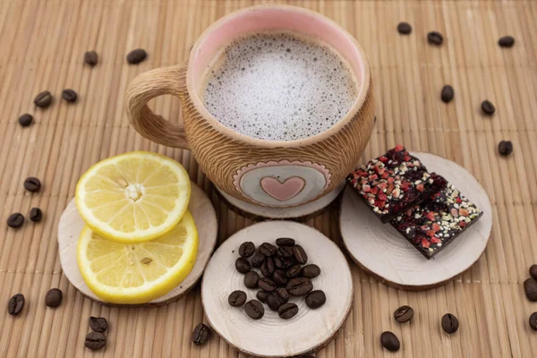 Ahşap tezgahta bir fincan kahve. Yakınlarda sarı sulu bir limon, arkaplanda meyveler ve kahve çekirdekleri serpiştirilmiş koyu çikolata bulunur. Yukarıdan görünüm.