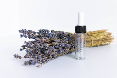 Beyaz arka planda kurutulmuş lavanta çiçekleri. Lavanta yağı. Pipetli şeffaf bir cam şişe buketin yanında duruyor.