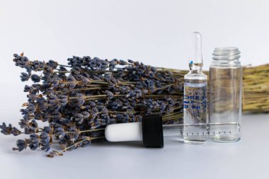 Beyaz arka planda kurutulmuş lavanta çiçekleri. Lavanta yağı. Pipetli şeffaf bir cam şişe buketin yanında duruyor.