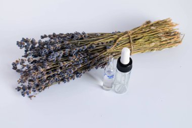 Beyaz arka planda kurutulmuş lavanta çiçekleri. Lavanta yağı. Pipetli şeffaf bir cam şişe buketin yanında duruyor.