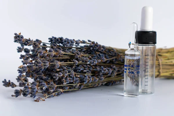 Beyaz arka planda kurutulmuş lavanta çiçekleri. Lavanta yağı. Pipetli şeffaf bir cam şişe buketin yanında duruyor.