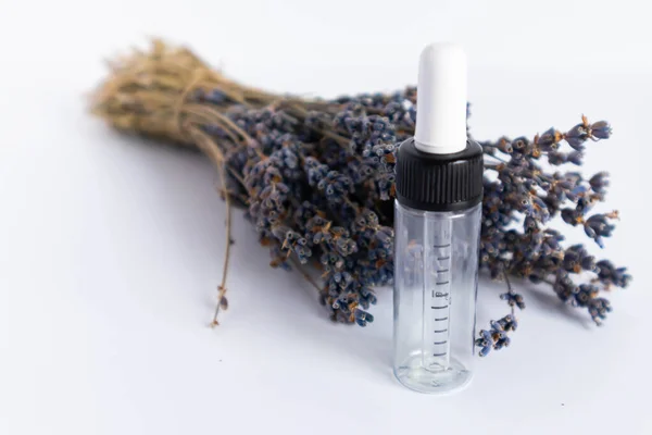 Beyaz arka planda kurutulmuş lavanta çiçekleri. Lavanta yağı. Pipetli şeffaf bir cam şişe buketin yanında duruyor..