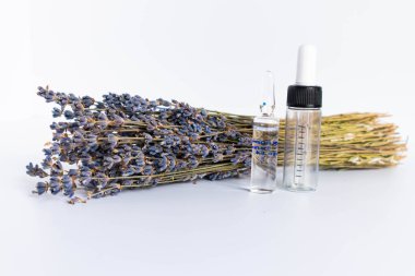 Beyaz arka planda kurutulmuş lavanta çiçekleri. Lavanta yağı. Pipetli şeffaf bir cam şişe buketin yanında duruyor..