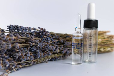 Beyaz arka planda kurutulmuş lavanta çiçekleri. Lavanta yağı. Pipetli şeffaf bir cam şişe buketin yanında duruyor..