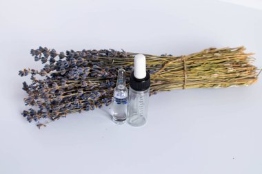 Beyaz arka planda kurutulmuş lavanta çiçekleri. Lavanta yağı. Pipetli şeffaf bir cam şişe buketin yanında duruyor..